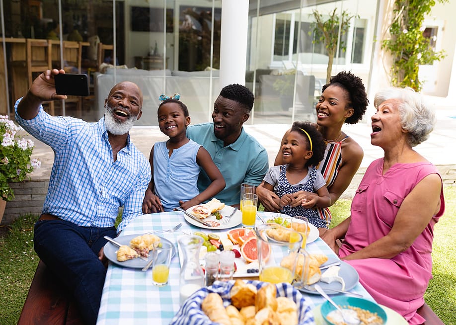 An older man smiling while taking selfie with his family at an outdoor dining table during brunch. — wavebreakmedia // Shutterstock