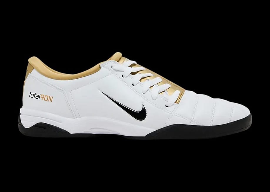 A pair of mostly white soccer shoes with a black Nike swoosh and a gold lining. — Courtesy of Sneakers.com