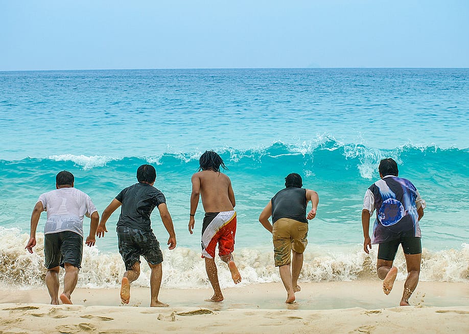 A group of five friends lined up to run towards a sea wave from the beach. — Lokal Brand // Shutterstock