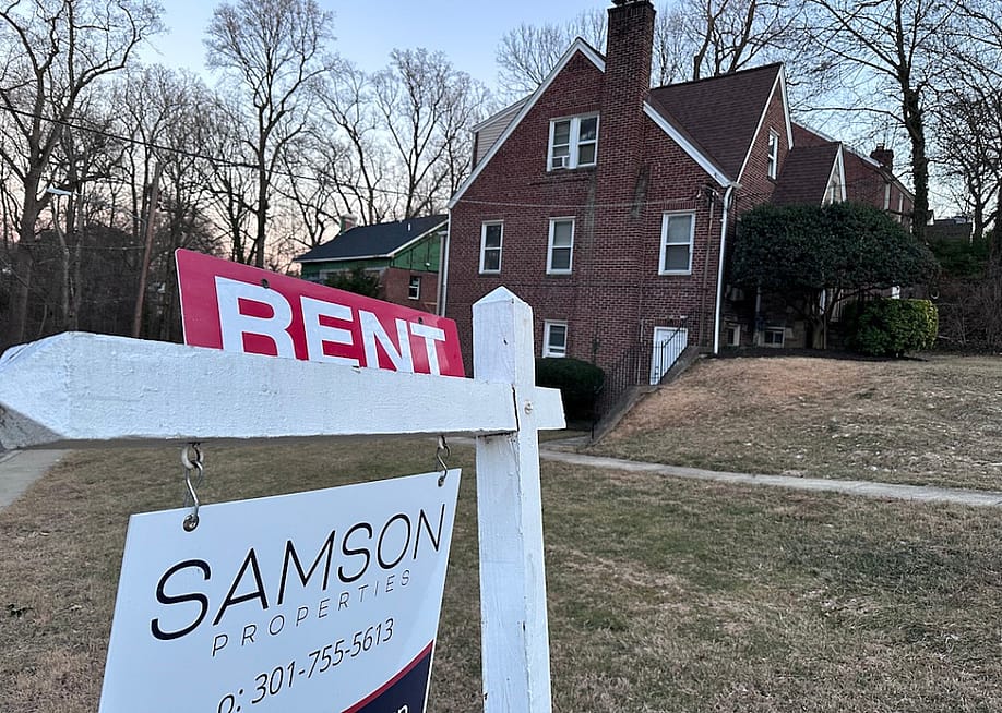 A sign in foreground lists a home for rent in Maryland with tall brick home in the background. Municipal rental registries are gaining attention as cities try to get a handle on who owns rental properties and where, both to better understand their housing landscape and to ensure rentals are safe for tenants. — Barbara Barrett // Stateline