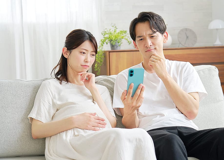 A pregnant wife and her husband curiously browsing information on a smartphone while sitting on a sofa at home. — siro46 // Shutterstock