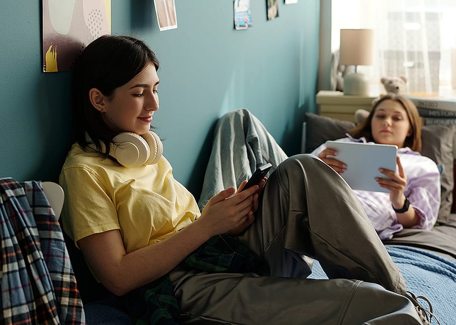 Two teenage girls relaxing on a bed and using smart devices to browse the internet. — Pressmaster // Shutterstock