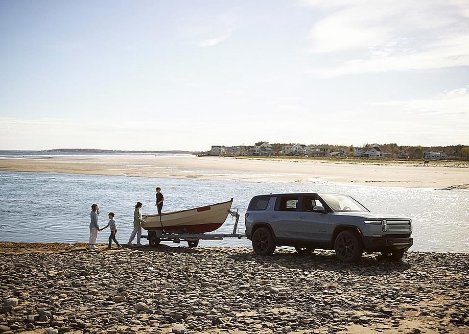 Rivian R1S towing a boat, parked near a body of water on a rocky dirt beach. A family of four plays near the boat. — Rivian