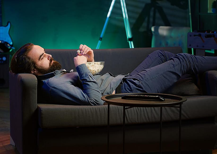 A bored man lying on couch and eating popcorn. — DC Studio // Shutterstock
