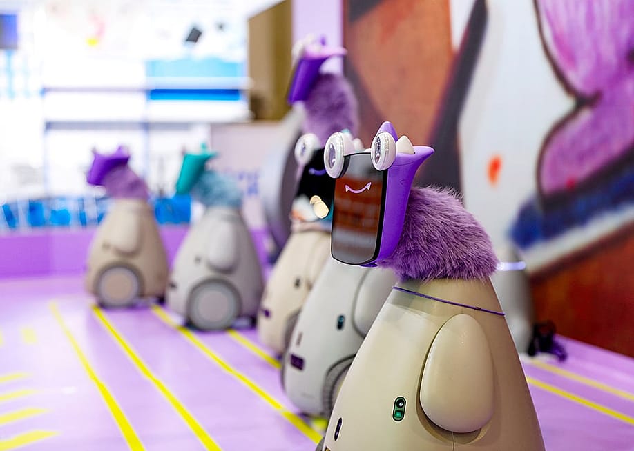 A row of 5 "robot pets" with playful googly eyes affixed on tablet faces and bright-colored faux fur necks on display at a convention. — Courtesy of OLLOBOT