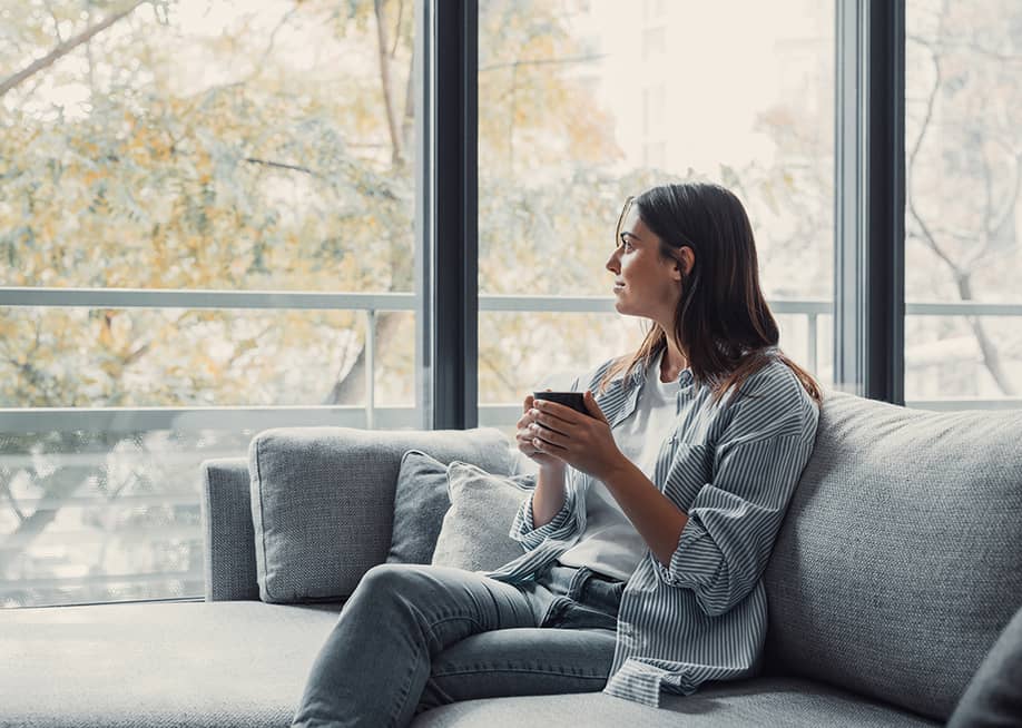 Young woman sitting on a sofa and having a cup of coffee while looking out the window at home. — PerfectWave // Shutterstock
