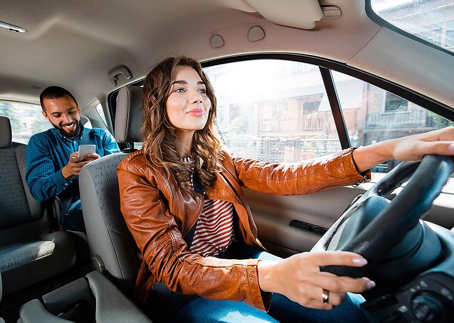 Wide shot of long-haired driver in brown leather jacket in the driver's seat with a passenger in backseat using phone. — Snapic_PhotoProduction // Shutterstock