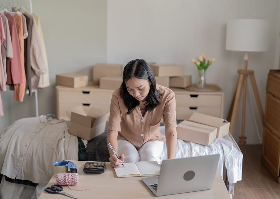 A woman writing in a notebook on a desk with a calculator, a laptop, scissors, tape and string, in a room with clothes hanging on a rack and cardboard boxes stacked on various surfaces. — Lee Charlie // Shutterstock