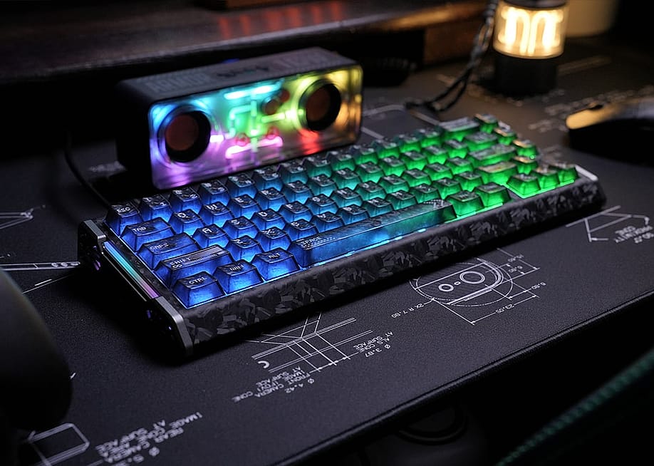 A blue-lit mechanical keyboard and a rainbow-colored speaker on a desk. — ATTACK SHARK
