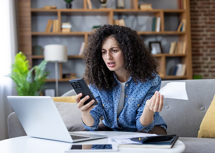 A young woman at home stressfully looking at her smartphone while calculating expenses. — voronaman // Shutterstock