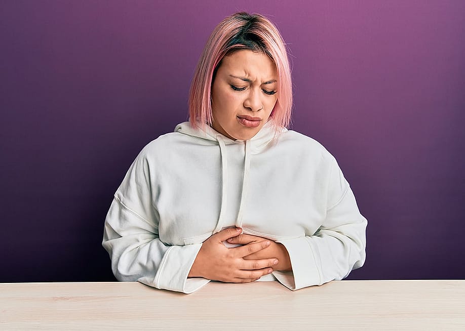A woman with pink hair sitting with her hands on her stomach, feeling unwell. — Krakenimages.com // Shutterstock