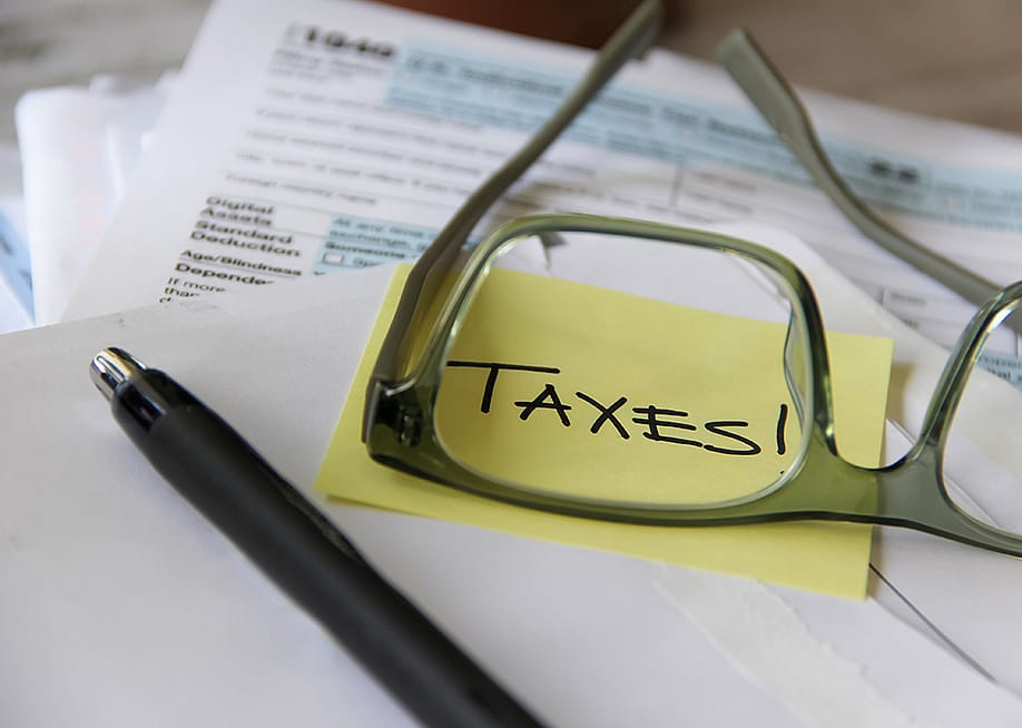 A low-angle view of tax documents and the word 'Taxes' written on a yellow sticky note viewed through eyeglasses. — MyLove4Art // Shutterstock
