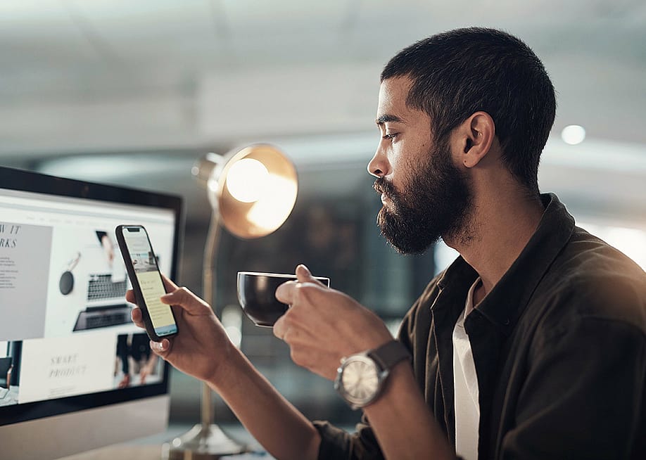 A man in a creative office checking social media on his phone while drinking a cup of coffee. — PeopleImages // Shutterstock