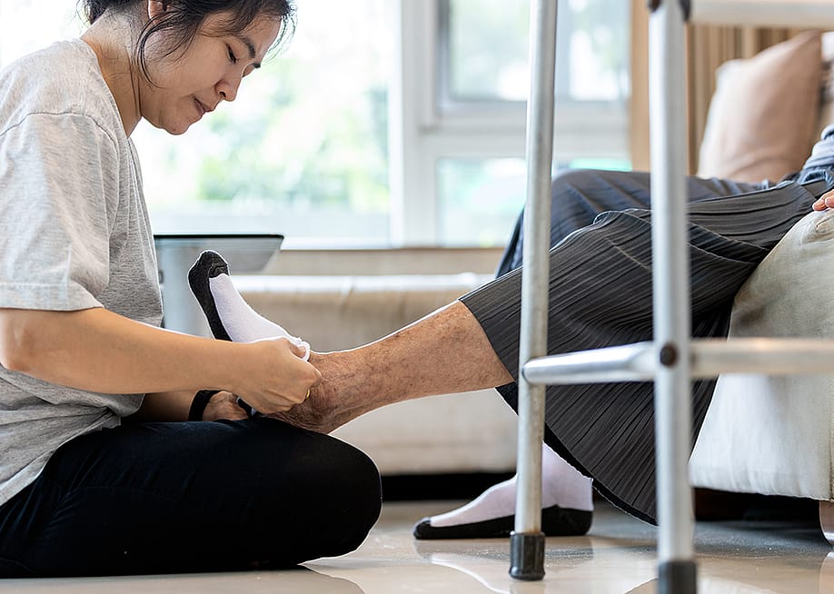 A female caregiver helping a senior person to wear socks at home. — CGN089 // Shutterstock
