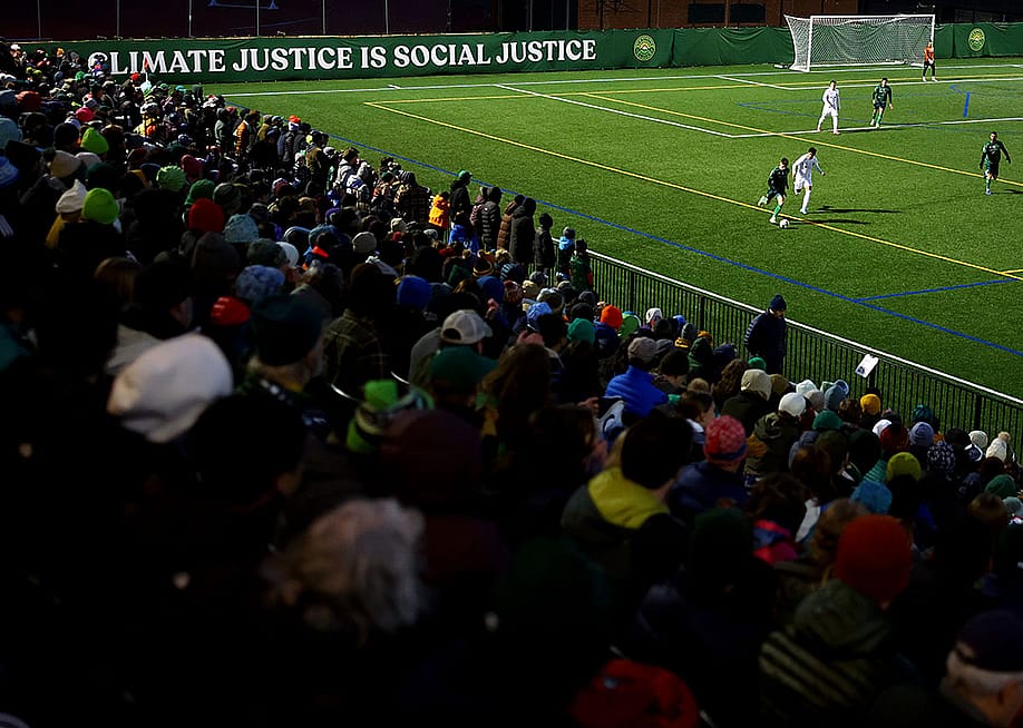 A view from the stands of the match with fans visible in the shadows on the bottom left and the green soccer pitch on the top right, with the words "Climate justice is social justice," displayed behind the goal during the between the Vermont Green FC and Lexington SC during the 2024 U.S. Open Cup at Virtue Field on March 19, 2024 in Burlington, Vermont. — Maddie Meyer // USSF / Getty Images for USSF