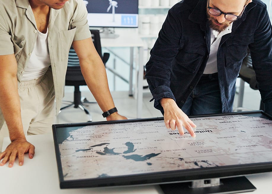 Two people looking at a U.S. map on a screen that is flat on a table in front of them. — PeopleImages // Shutterstock