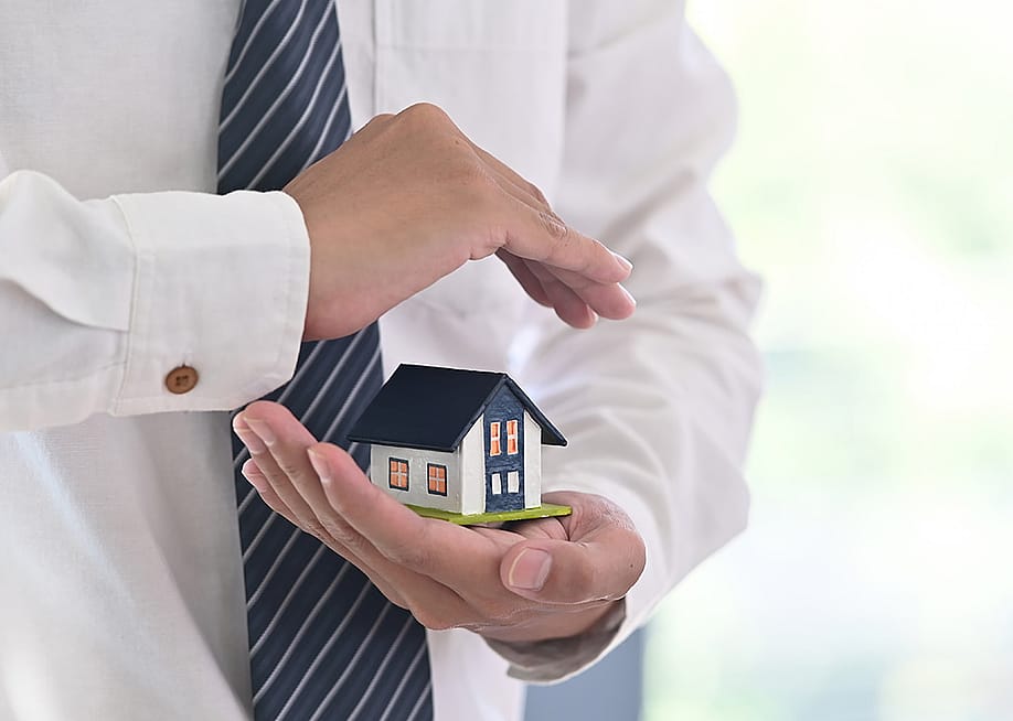 A real estate agent holding a small figure of a house in a protective gesture as a concept of insurance. — Prathankarnpap // Shutterstock