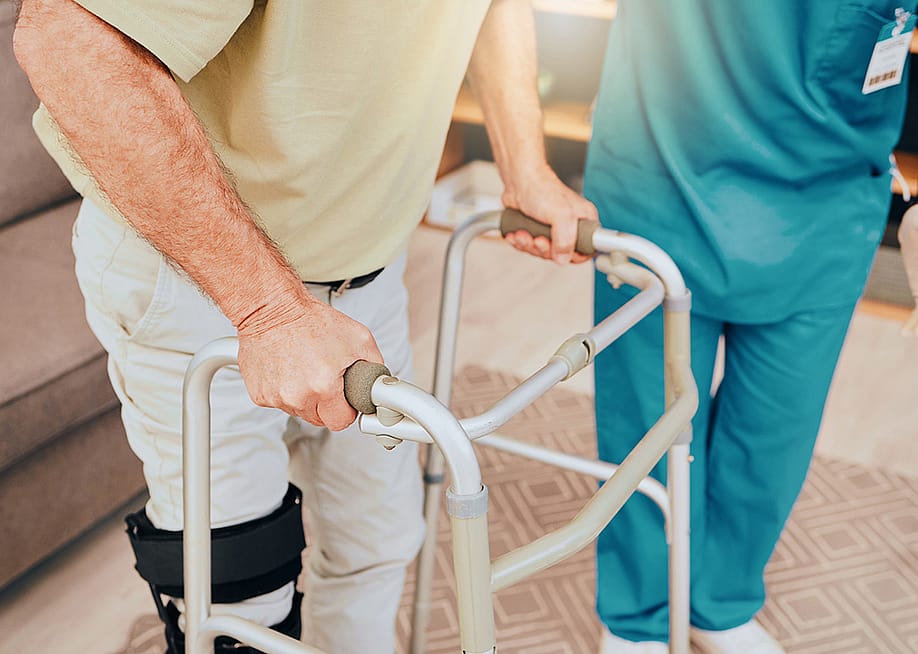 A senior man using a walker during a physiotherapy session assisted by a caregiver. — PeopleImages // Shutterstock