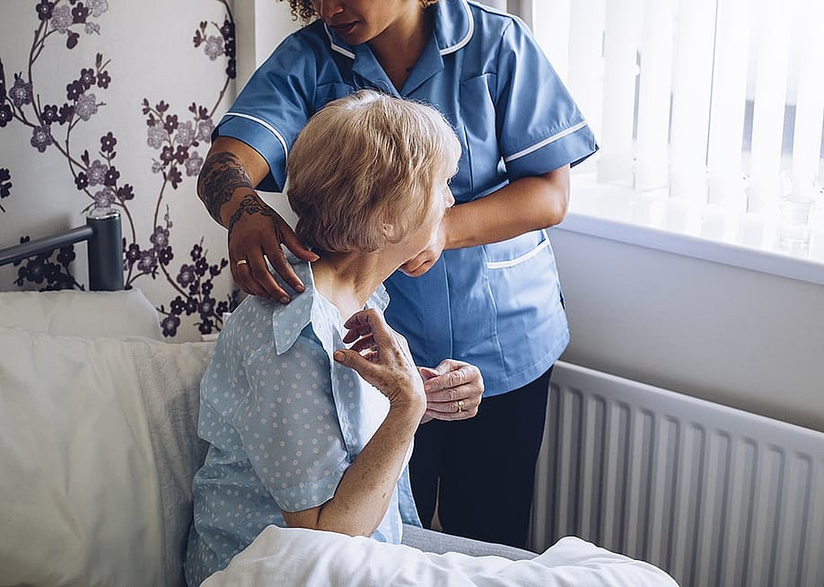 A home caregiver helping a senior woman get dressed in bed. — DGLimages // Shutterstock