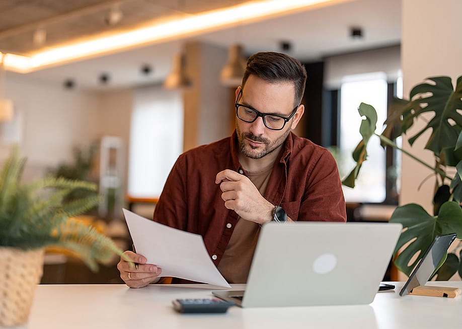 A person looking at a paper and using a laptop in a home office. — Dorde Krstic // Shutterstock