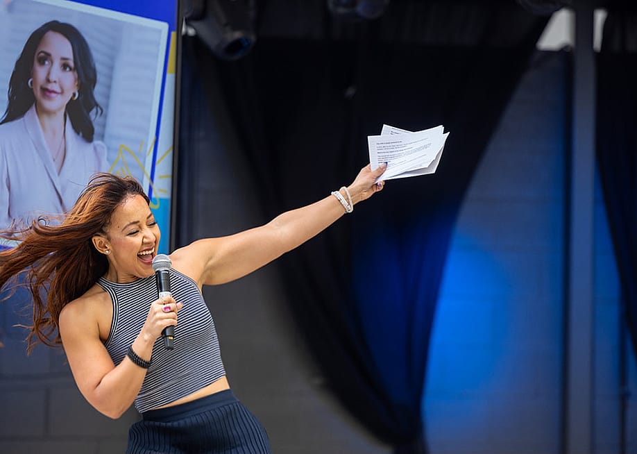 Julia Collins who is the founder and CEO of Planet FWD, speaks onstage with one hand holding a microphone and the other arm raised, her portrait is visible on a screen in the background. — Chloe Jackman Photography