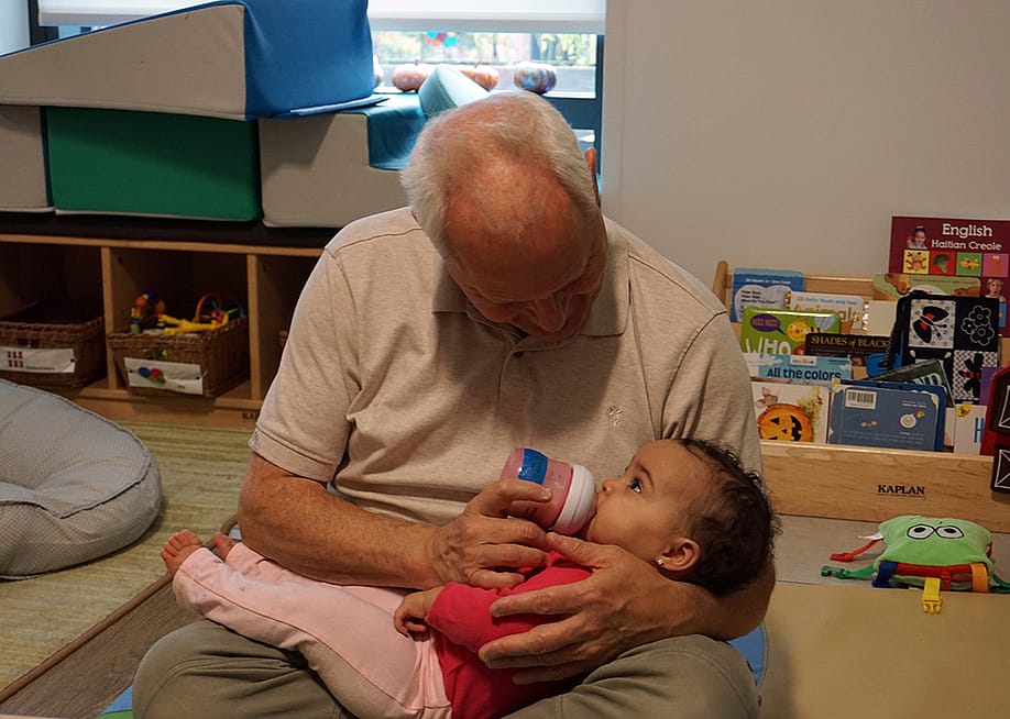 Teacher Herb Hickey holds a baby while she drinks from a bottle. — Jackie Mader for The Hechinger Report