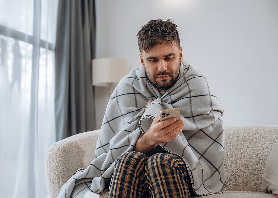 A sick man at home wrapped in blanket and looking up information on his phone. — Standret // Shutterstock
