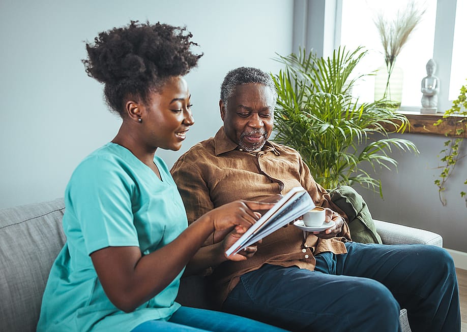 A black mid-adult nurse attentively aids an elderly black male with his reading material. — Dragana Gordic // Shutterstock