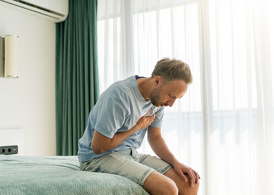 A man sitting on his bed clutching chest due to pain. — DedMityay // Shutterstock