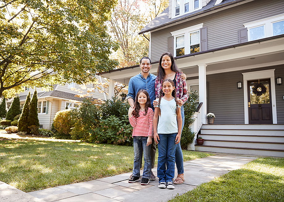 A family of four happily standing in front of their home for a photo. — Monkey Business Images // Shutterstock