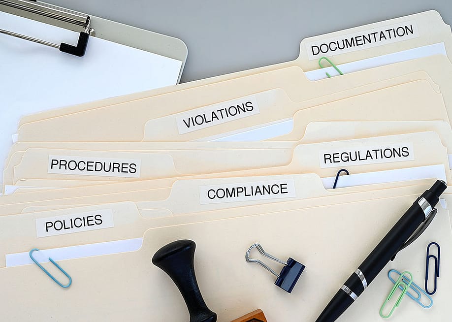 Folders of compliance and policy documents on a desk. — MargJohnsonVA // Shutterstock