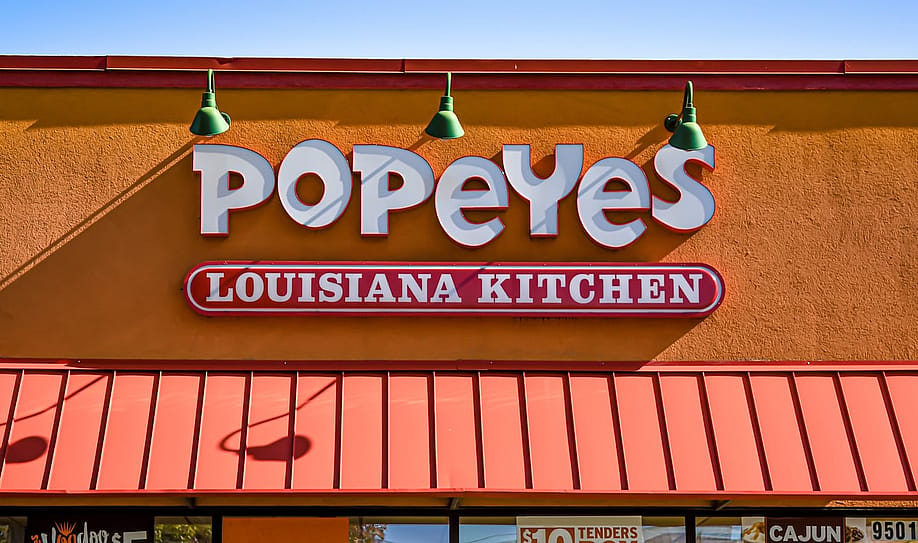 orizontal, medium closeup of "Popeyes" Louisiana Kitchen exterior facade brand and logo signage in white letters on a bright sunny day. — Bruce VanLoon // Shutterstock