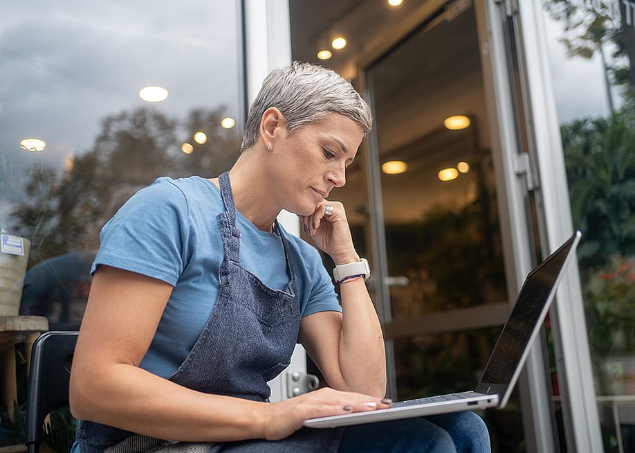 A female small business owner by the entrance of her store reviewing information on a laptop. — Nenad Cavoski // Shutterstock