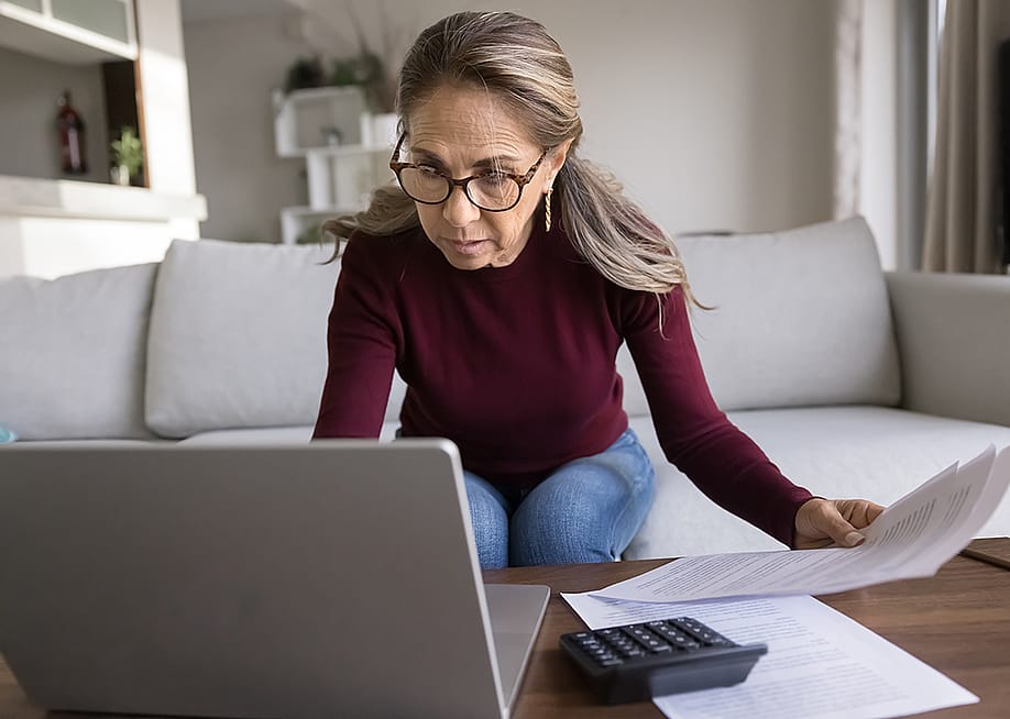 A focused senior woman holding financial documents and browsing information on a laptop. — fizkes // Shutterstock