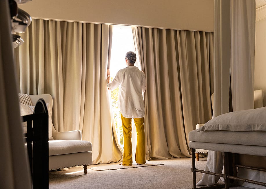 A senior woman standing by the large window of a cozy living room. — wavebreakmedia // Shutterstock