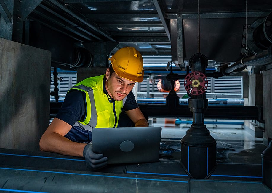 An industrial engineer viewing a laptop while checking an HVAC pipeline system. — CandyRetriever // Shutterstock