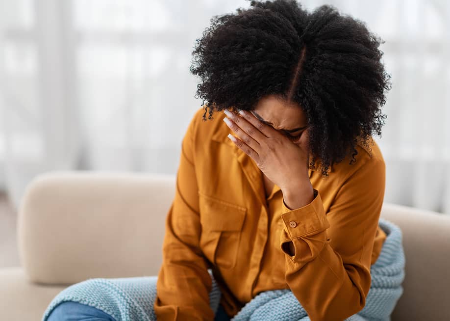 A young black woman sitting on a sofa holding her due to stress. — Prostock-studio // Shutterstoc