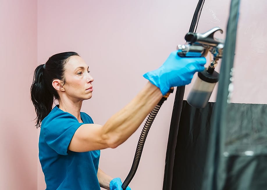 A beauty technician applying spray tan to a client inside a black booth. — LMeisy // Shutterstock