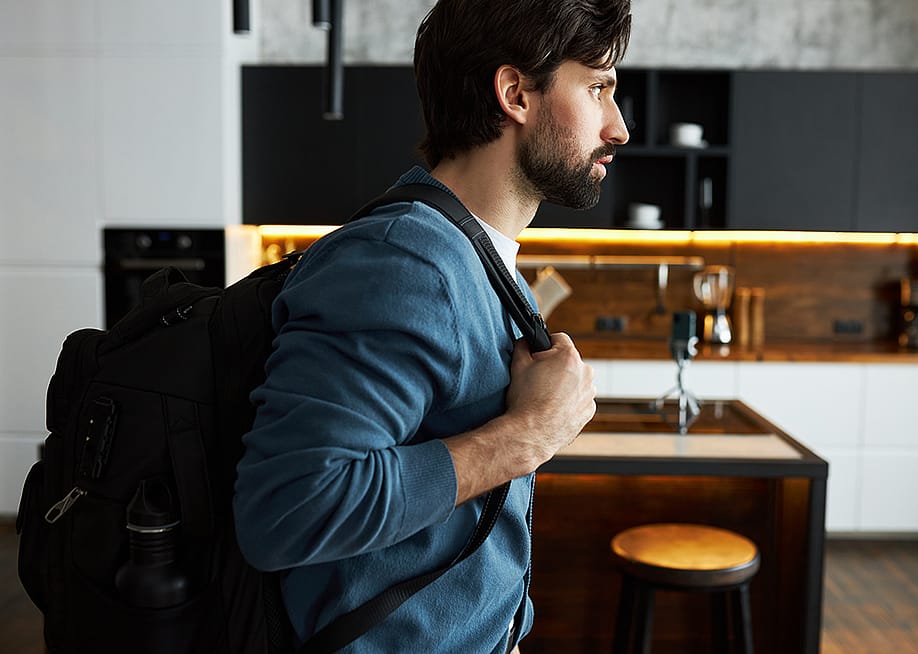 Man at home in a blue cardigan and putting on a black backpack preparing for a trip. — Anatoliy Karlyuk // Shutterstock