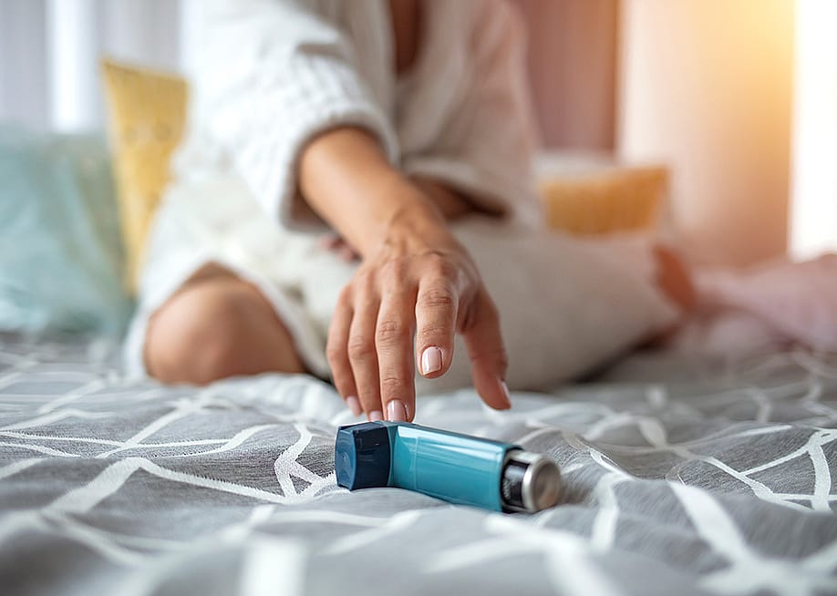 Woman sitting on a bed reaching for her inhaler. — Dragana Gordic // Shutterstock