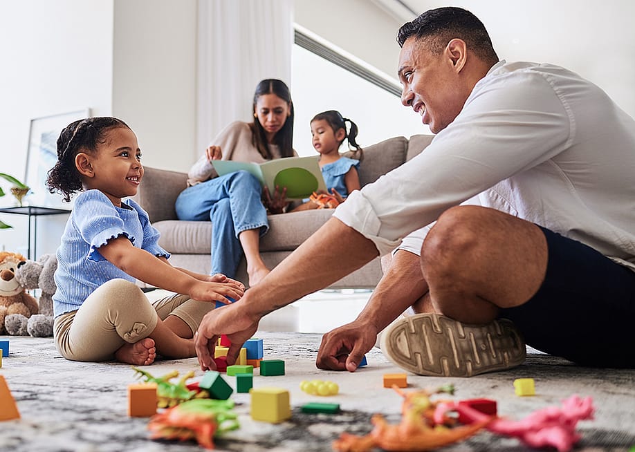 Parents playing with and reading a book to their young daughters at home. — PeopleImages // Shutterstock