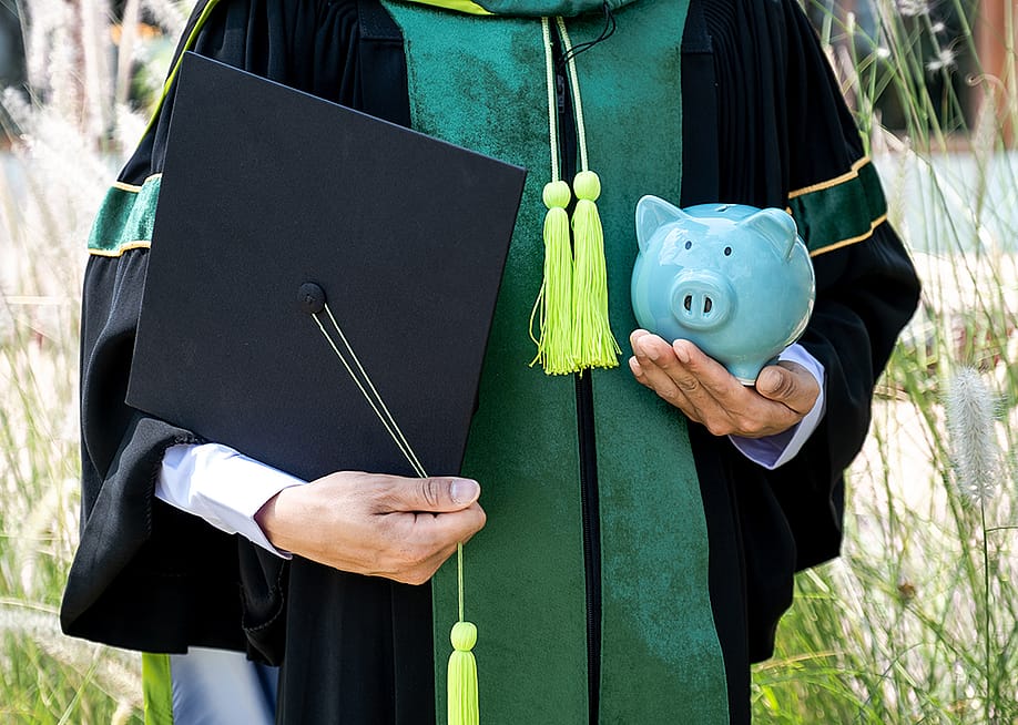 A person wearing a bachelor uniform holding a cap and a piggy bank. — Inthon Maitrisamphan // Shutterstock