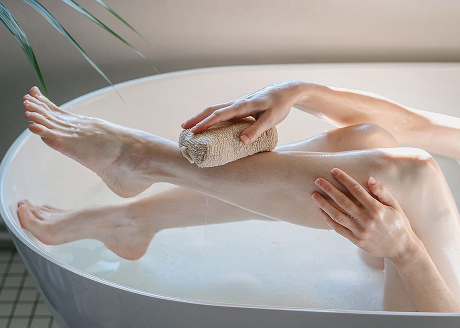 A cropped view of a person washing her legs in a bath tub with a body sponge. — brizmaker // Shutterstock