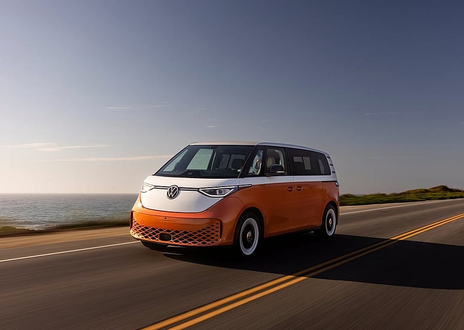 Candy White over Energetic Orange 2025 Volkswagen ID Buzz driving on road next to ocean. — Volkswagen