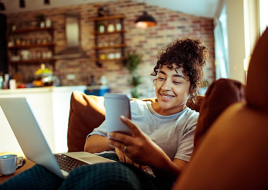 A young woman at home smiling at her phone while relaxing on a couch. — Davor Geber // Shutterstock