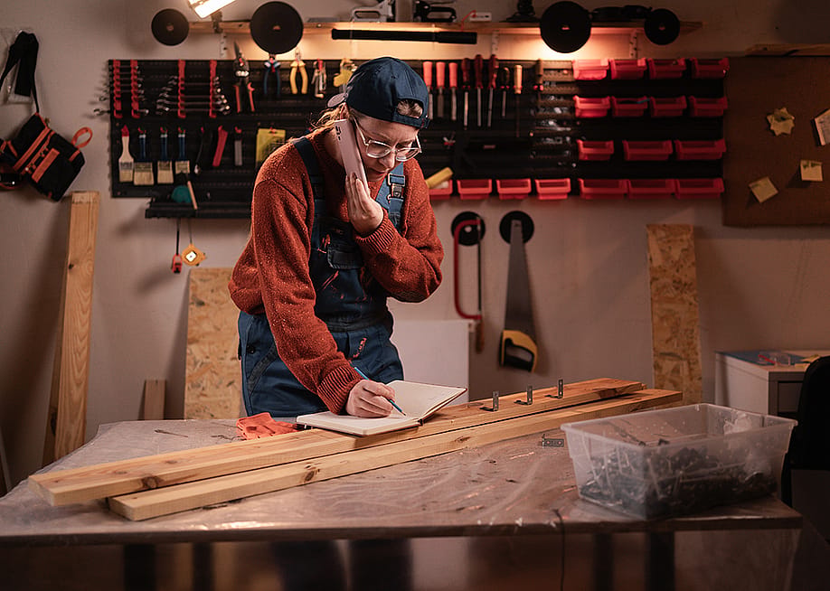 A female carpenter writing down notes while talking on the phone in a workshop. — Lysenko Andrii // Shutterstock