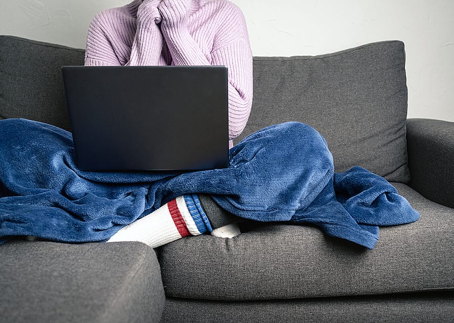 Person wearing a knitted sweater and socks with a blanket on while working and sitting on a sofa. — Ripio // Shutterstock