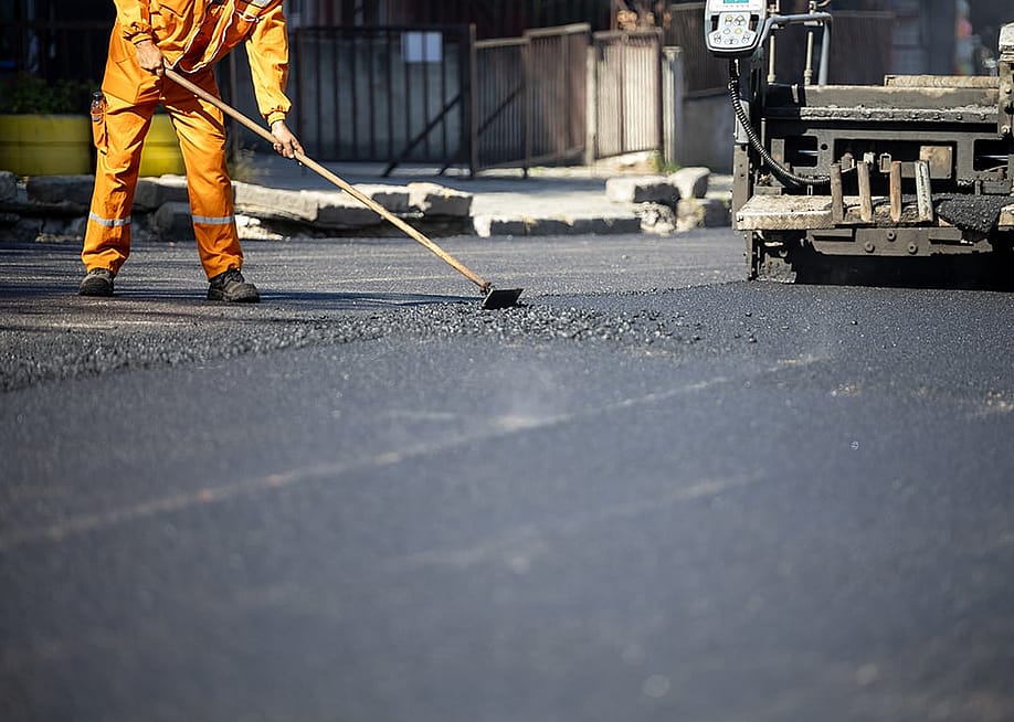 A construction worker in an orange uniform spreads freshly poured asphalt on a road surface. — zoff // Shutterstock