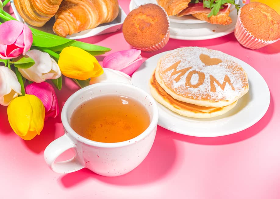 A pink-themed Mother's Day brunch meal featuring a bouquet of flowers, a cup of tea, and pancakes. — Rimma Bondarenko // Shutterstock
