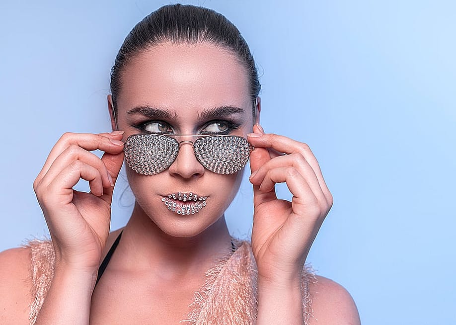 A female model wearing a rhinestone sunglasses and with rhinestones makeup on her lips. — TinaGreen // Shutterstock